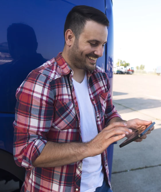 Uśmiechnięty mężczyzna w kratowanej koszuli korzystający z tabletu oparty o bok niebieskiego pojazdu na zewnątrz