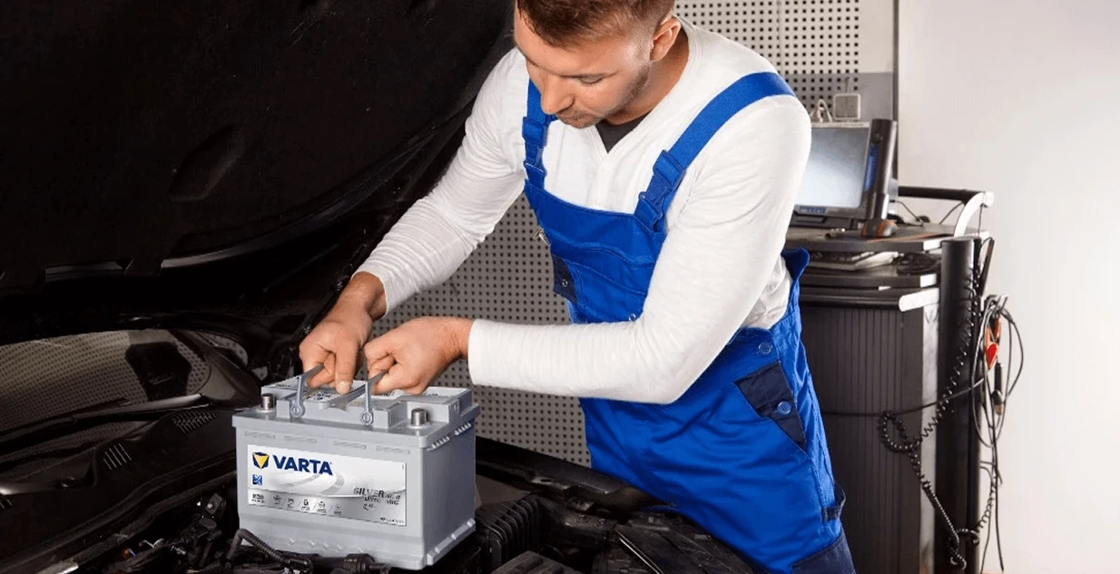Mechanik w niebieskim kombinezonie montuje akumulator VARTA pod maską auta, w tle sprzęt diagnostyczny