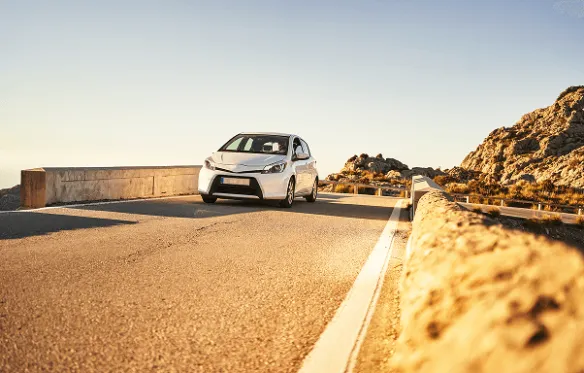 Bijeli automobil na cesti uz more Bijeli automobil vozi po cesti uz stijene i more u zlatnom svjetlu zalaska sunca.