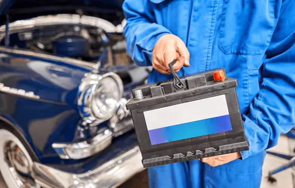 Mužjak u plavom kombinezonu drži automobilsku bateriju pored klasičnog automobila.