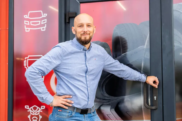 Vīrietis stāv pie automobiļu servisa durvīm ar riepu attēlu fonā.