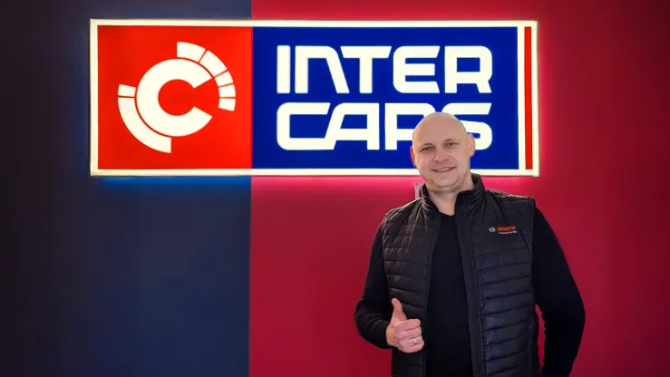 Inter Cars zīmola logo uz sarkanas un zilas fona, cilvēks pozē ar pirkstu uz augšu.