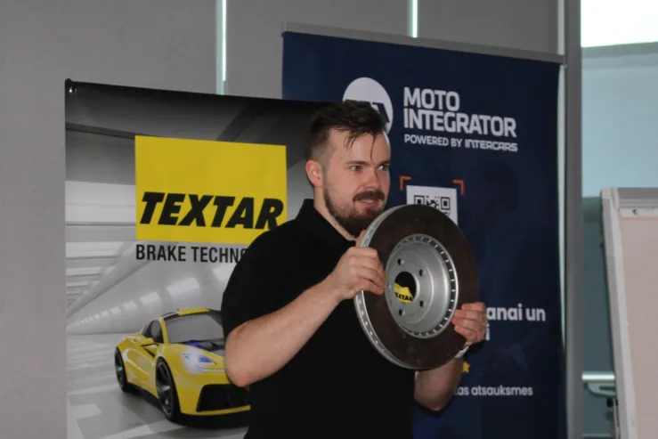 Vīrietis demonstrē Texar bremžu disku prezentācijā ar automobiļa attēlu fonā.
