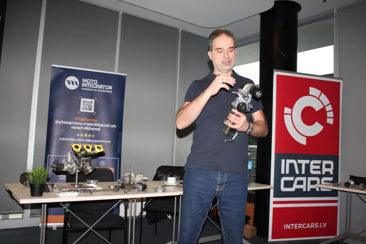 Vīrietis demonstrē auto detaļu, fonā Moto Integrator un Inter Cars reklāmas stendi.