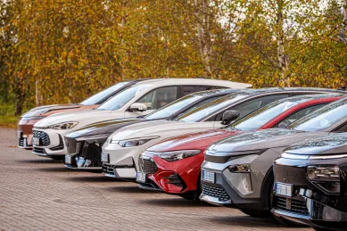 Rinda ar dažādiem automobiļiem, kas novietoti uz bruģēta ceļa, ar dzeltenām lapām fonā.