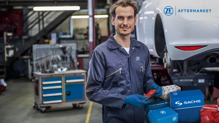 Mechanik w niebieskim kombinezonie trzyma części samochodowe w warsztacie, z narzędziami w tle.