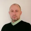 Mężczyzna w czarnym swetrze i zielonej koszuli, stoi na neutralnym tle.