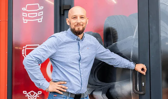 Vīrietis stāv pie automobiļu servisa durvīm ar riepām fonā.