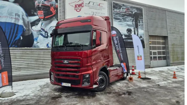 Czerwony ciężarówka Ford F-MAX GEN2 zaparkowana przed budynkiem, z flagami i plakatami w tle.