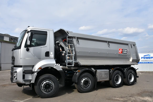 A silver dump truck parked, featuring a ladder and a large cargo bed, with a clear sky in the background.
