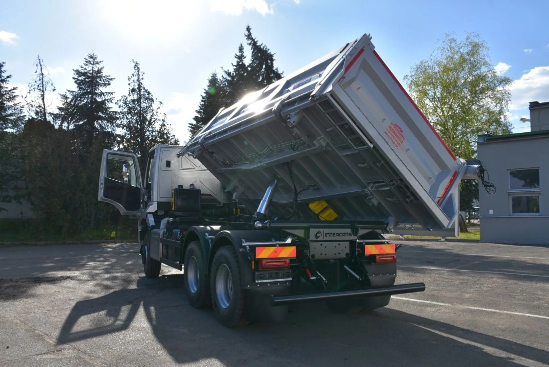 Wywrotka z podniesioną skrzynią, prezentująca mechanizm załadunku, na tle drzew i budynku.