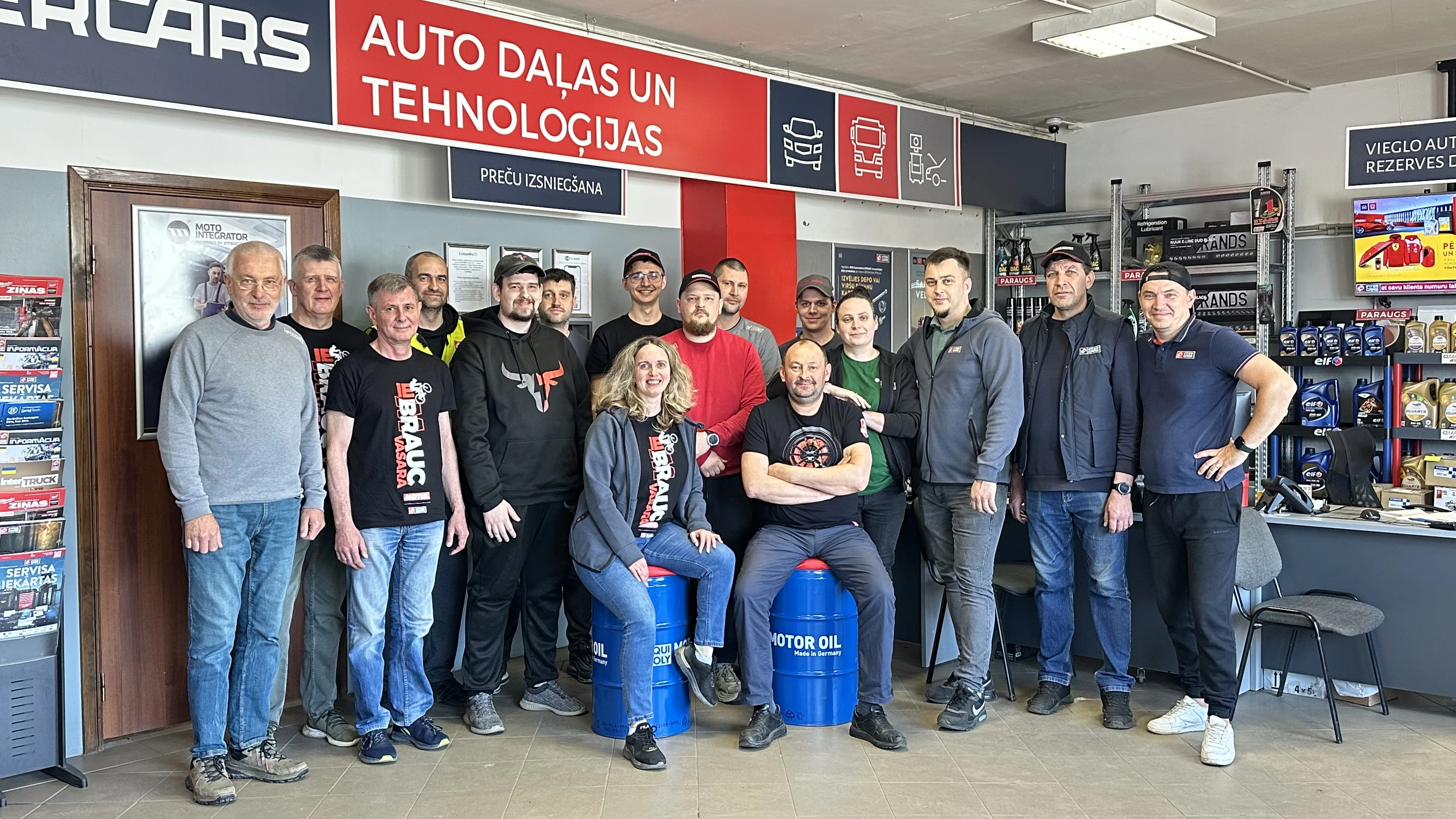 Auto detaļu un tehnoloģiju veikala komanda Darbinieku grupa, kas pozē auto detaļu veikalā ar dažādiem produktiem un izkārtnēm fonā.