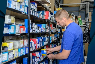Pracownik magazynu w niebieskiej koszuli ogląda przedmioty na półkach wypełnionych kolorowymi pudełkami i częściami.