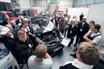 Grupa ludzi skupiona wokół otwartego silnika samochodu w warsztacie mechanicznym, słuchająca wyjaśnień prowadzącego, który wskazuje na szczegóły techniczne pojazdu