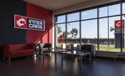 Nowoczesne wnętrze poczekalni z dużymi oknami, wyposażone w czerwoną sofę i stolik kawowy, z logo "Inter Cars" na ścianie