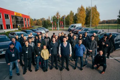 DHL darbinieku grupa stāv pie automašīnām un biroja ēkas.